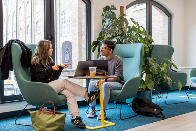 Two people are seated in modern teal armchairs, engaged in conversation in a stylish office space. A laptop and iced coffee are on the table between them. Large windows and green plants create a bright, inviting atmosphere.