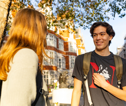 Two people with backpacks are talking outdoors in a sunlit area. One is a woman with long hair, and the other is a man wearing a T-shirt with a dog graphic. They are standing near a large brick building surrounded by trees.