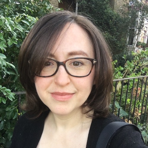 A person with shoulder-length brown hair and glasses smiles while standing outdoors. Greenery and a metal fence are visible in the background.