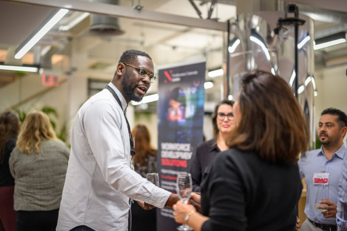 A group of people socializing at an indoor event. A man in a white shirt and dark vest is shaking hands with a woman in black. A blurred banner and other attendees are in the background. The setting appears to be an office or conference space.