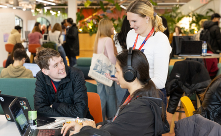 A group of students in a modern study area, with one standing and talking to two others seated with laptops. The space is lively with greenery and people in the background. They appear engaged and focused.