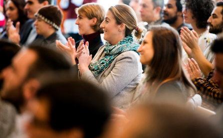 A group of people seated in an audience clapping. The focus is on a woman in the center wearing a blue scarf and light-colored jacket, surrounded by others who are also applauding. The background is blurred.