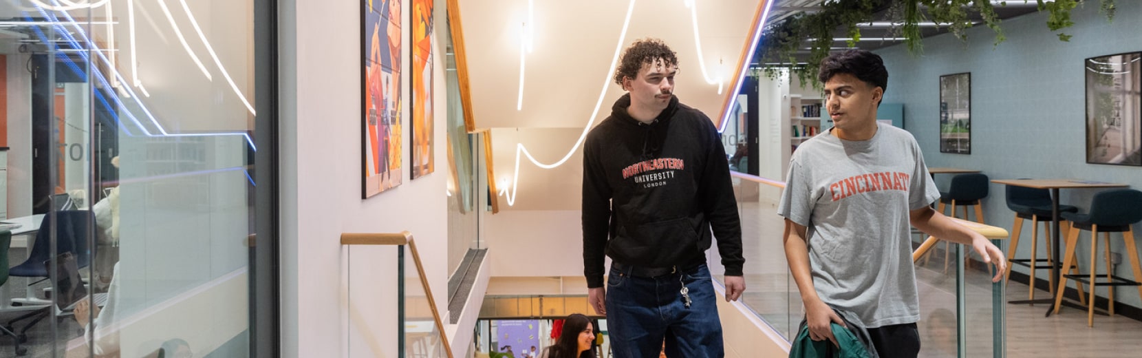 Two people walk through a modern building with plants and artwork on display. One wears a black hoodie, the other a gray T-shirt. The space is brightly lit, with a staircase and seating area visible.