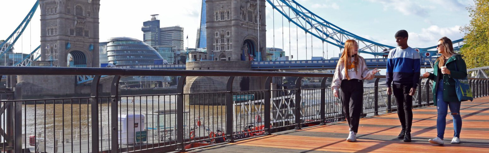 Three people walk along a riverside path near Tower Bridge in London. The bridge's iconic towers and suspension cables are visible in the background, alongside modern buildings and the Thames River.