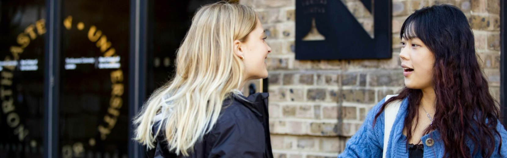 Two students conversing outside a university building. One has long blonde hair and is dressed in a dark jacket, while the other has long dark hair and wears a blue sweater. The university sign is visible on the brick wall behind them.