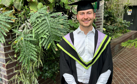 A graduate wearing a cap and gown with green and purple trim stands smiling in front of a leafy brick wall.