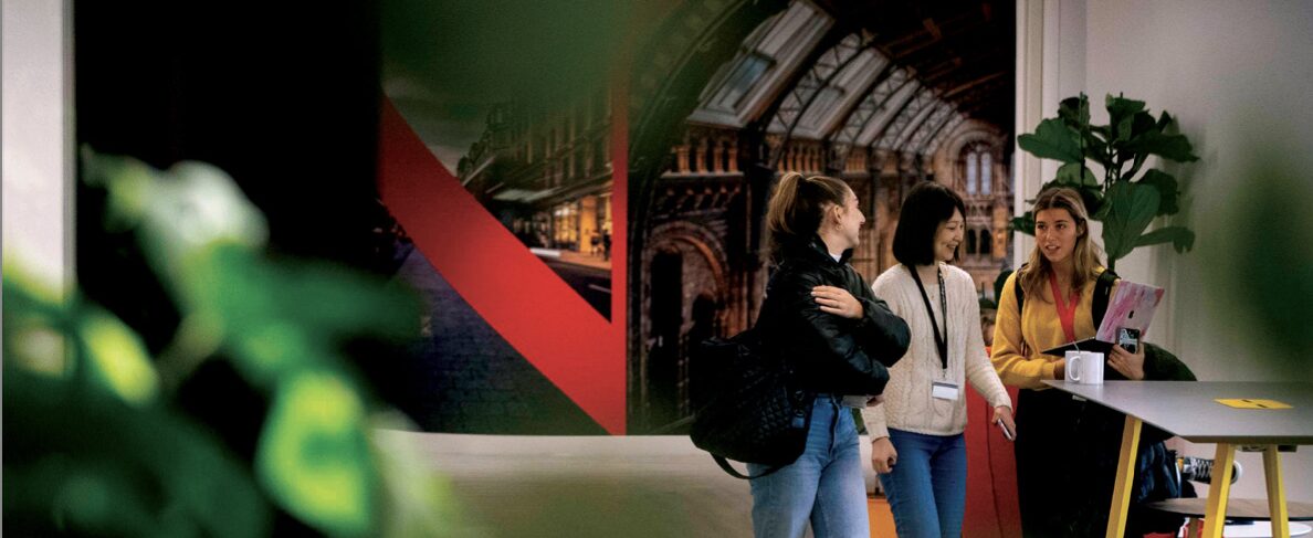 Three people stand in a hallway, chatting and holding notebooks. They are near a large mural depicting an architectural scene. Green plants are in the foreground, partially obscuring the view.