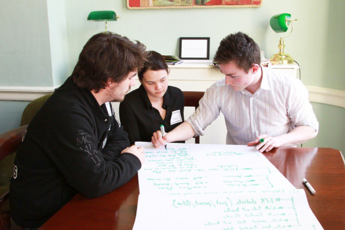 Three people are seated around a table, collaborating and writing on a large sheet of paper. Two are men, and one is a woman. They appear focused and engaged in a discussion, possibly brainstorming ideas. The setting looks like a meeting room.