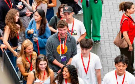 A group of people is gathered outdoors, many wearing red lanyards. Some are smiling and chatting. There are a mixture of men and women, dressed casually. The background includes a few spectators and a person in bright green pants.