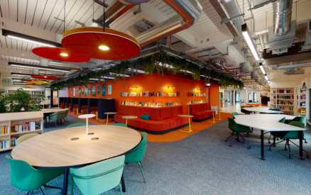 A modern library interior with colorful round tables and green chairs. Bookshelves line the orange walls, and greenery hangs from the ceiling. The space is well-lit with large windows and industrial ceiling features.