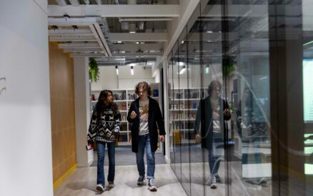 Two people walking down a hallway with glass walls. The person on the left is holding a phone and wearing a patterned sweater, while the person on the right is wearing a coat and jeans. Bookshelves are visible in the background.