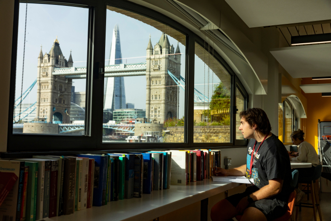 A man sits at a table in a library, reading a book. Through the large windows, London’s Tower Bridge and The Shard are visible. Books line the window ledge, and another person is working in the background.