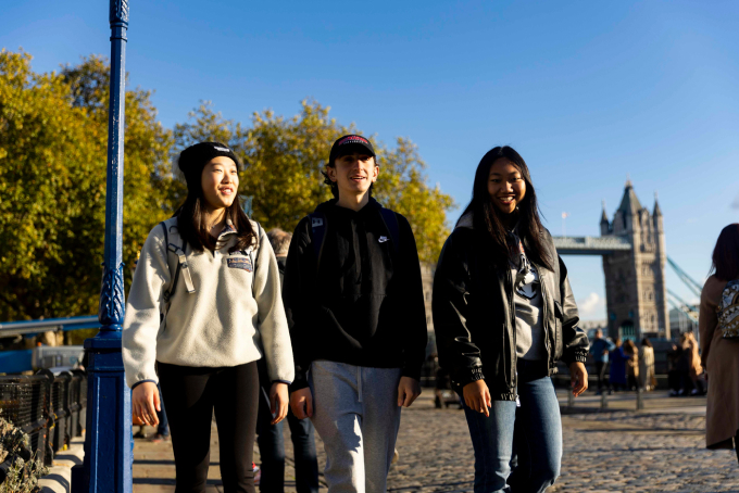 Three people walking and smiling in front of Tower Bridge, London. The sunny day highlights the autumn foliage in the background. They are dressed casually in jackets and hoodies.