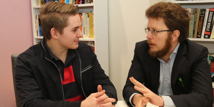 Two men are engaged in conversation in a room with bookshelves. The man on the left has short hair and wears a black jacket, while the man on the right has curly hair, glasses, and a beard, wearing a suit. They appear to be discussing something intently.