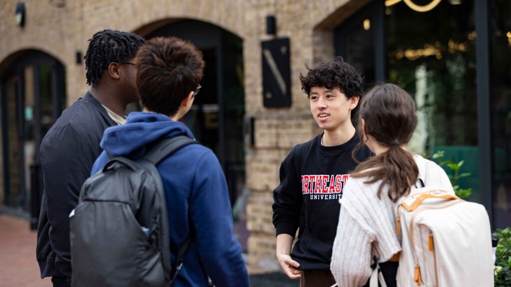 Four young adults stand outside a brick building, engaged in conversation. One wears a "Northeastern University" sweatshirt. They all carry backpacks, suggesting a college campus setting.