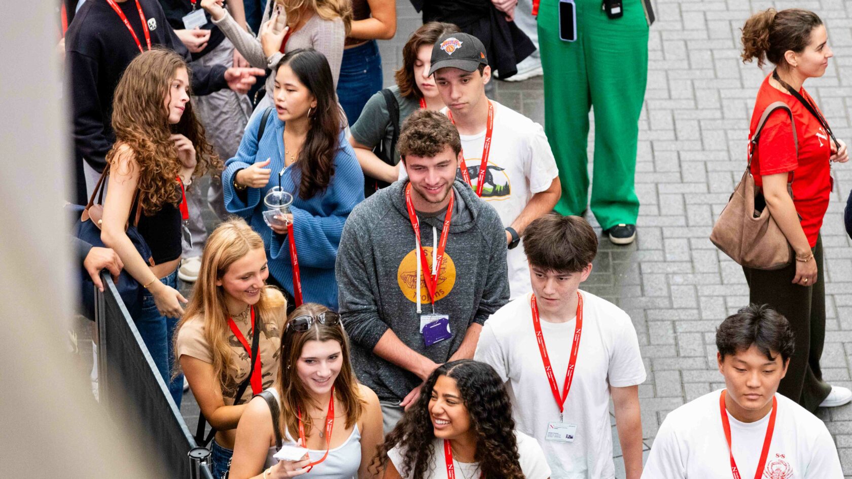 A diverse group of young people wearing red lanyards walk together outdoors on a paved pathway. They appear engaged and are dressed casually, holding drinks or phones, with some smiling. The setting seems to be a social or event gathering.