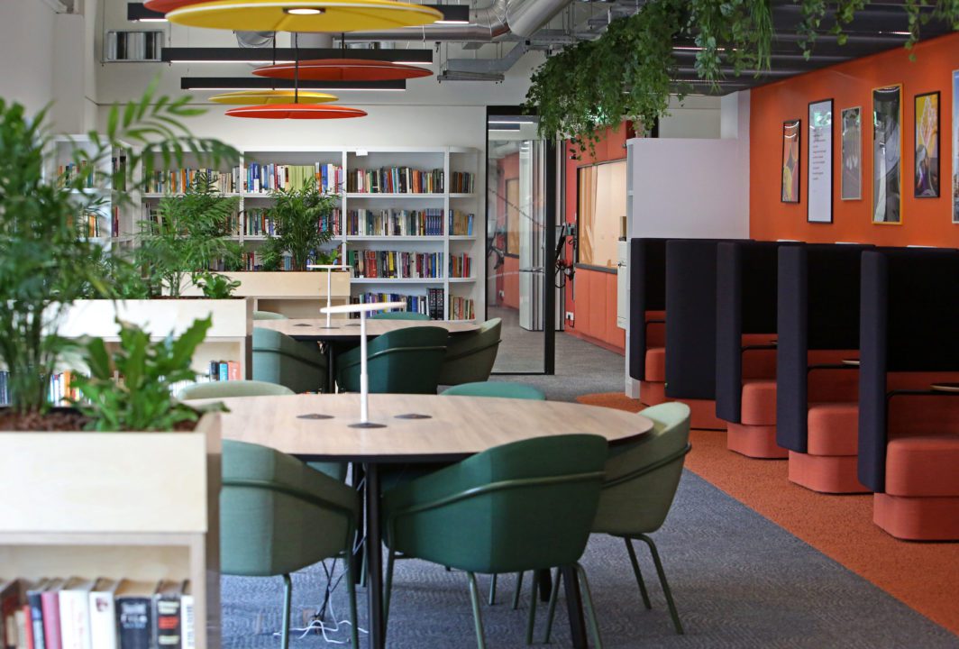 Devon House. A modern library interior featuring round tables with green chairs, bookshelves filled with books, and a wall with colorful framed posters. Ceiling lights with orange and yellow shades hang above, and greenery adds a fresh touch.