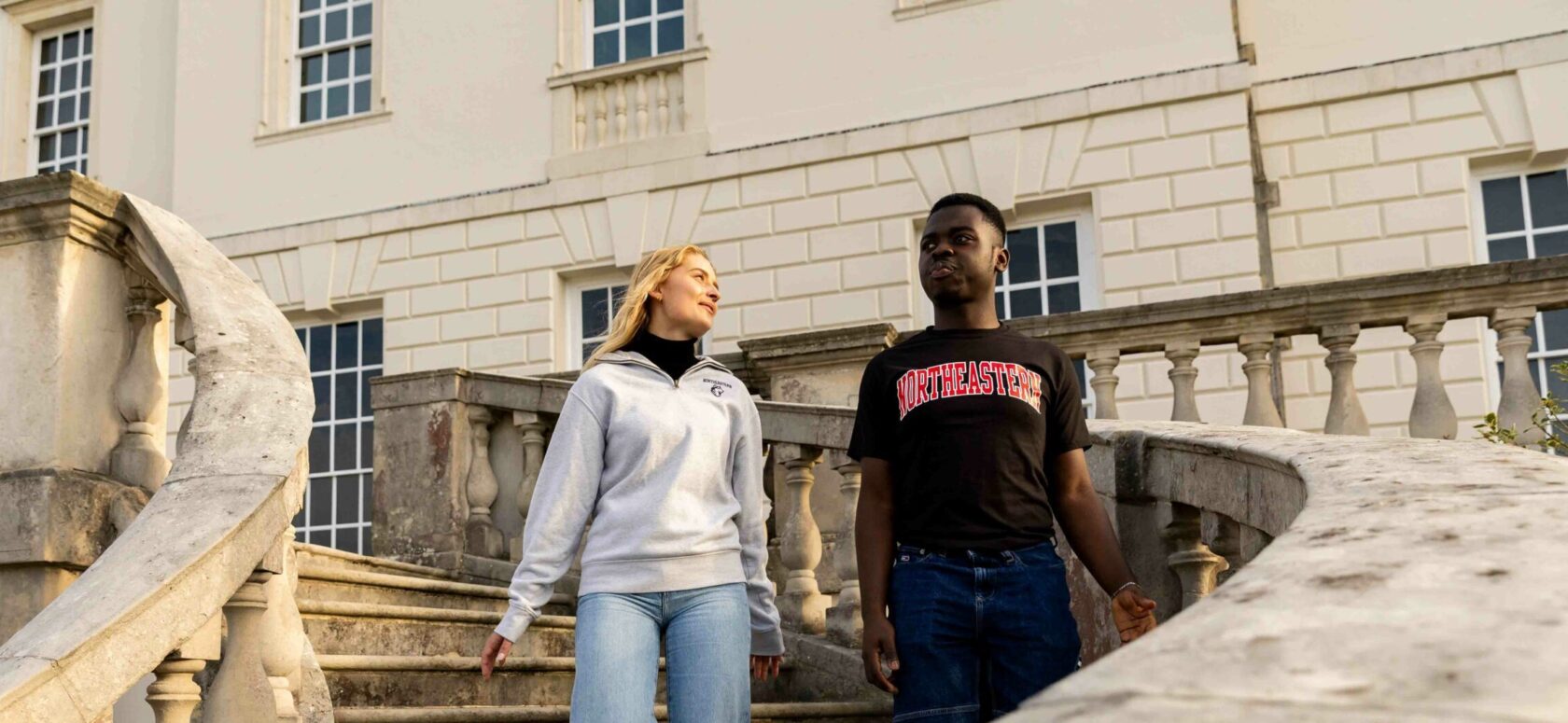 Two people are walking down a stone staircase in front of a large building with white walls and large windows. One wears a light gray sweater, and the other wears a black T-shirt with red text.