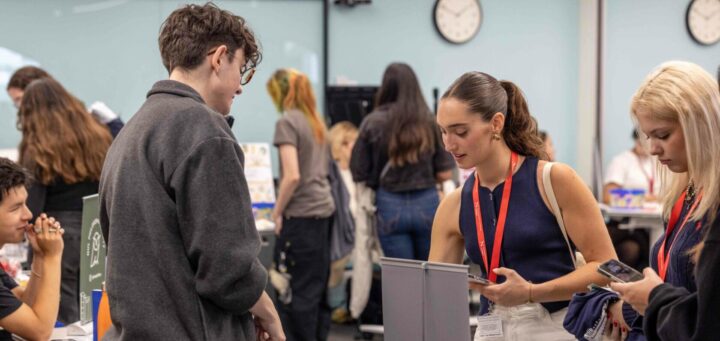 A group of people interacting at a busy event. One woman with a red lanyard assists a man with glasses at a table. Others nearby are engaged with their phones or talking. Clocks on the wall indicate different times, suggesting an international atmosphere.