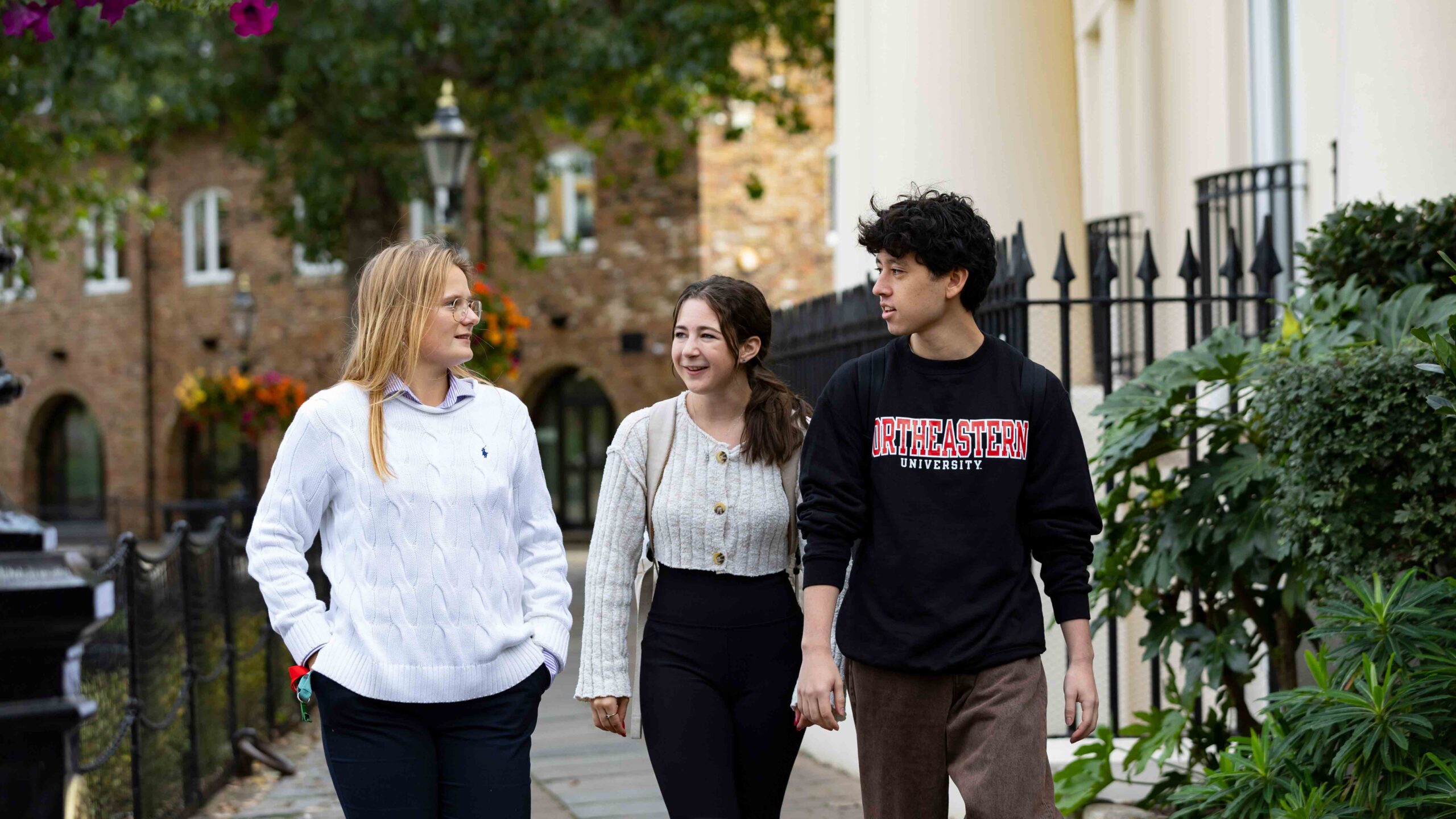 Three people walking down a sidewalk beside a building with ornate fencing and greenery. One wears a white sweater, another a beige cardigan, and the third a black sweatshirt with u0022Northeastern Universityu0022 written on it. Trees and a lamppost are in the background.
