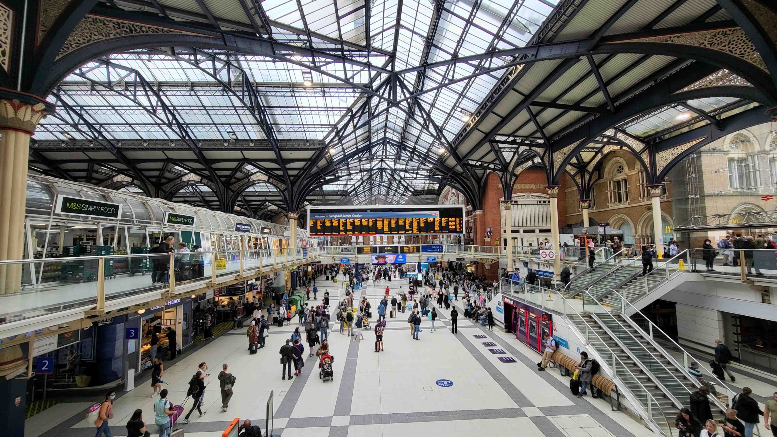 A bustling railway station with a large glass roof and ornate ironwork. People walk across the concourse, and a digital departure board displays train information. Shops and staircases line the sides, with escalators leading to an upper level.