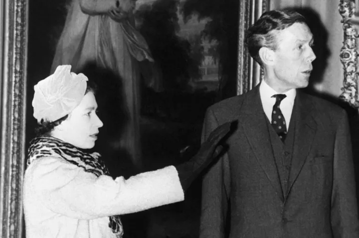 A woman in a patterned coat and hat reaches out to gesture toward a man in a suit and tie. They are both standing in an ornate room with a large framed painting in the background. The man is looking to the side.