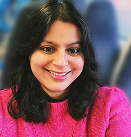 A person with shoulder-length dark hair is smiling, wearing a bright pink sweater. The background is blurred, suggesting an indoor setting.