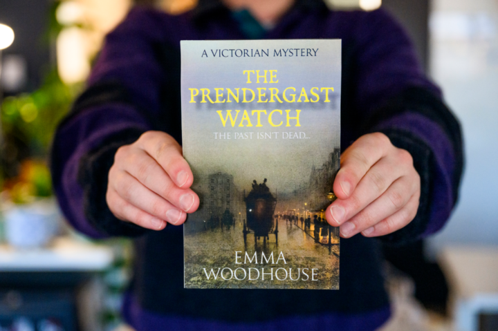 Person holding a book titled "The Prendergast Watch" by Emma Woodhouse. The cover shows a vintage street scene with a horse-drawn carriage and fog. Words "A Victorian Mystery" and "The past isn't dead..." are above the title.