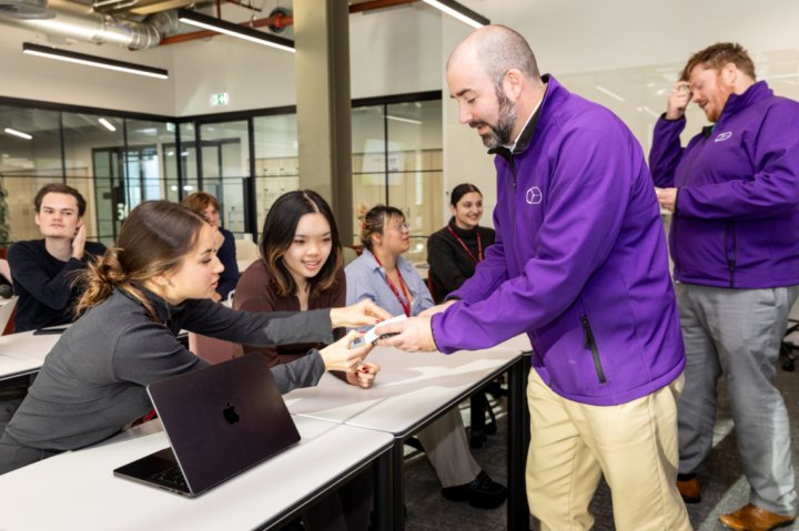 A man in a purple jacket hands a card to a woman in a classroom setting. Several people sit at tables with laptops. Another person in a purple jacket is in the background, engaged with others. The room has a modern design with glass walls.