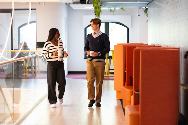 A woman and a man are walking and talking in a modern office hallway. She holds a phone, and he holds a cup. They are passing by orange booths and large windows. Green plants hang from the ceiling.