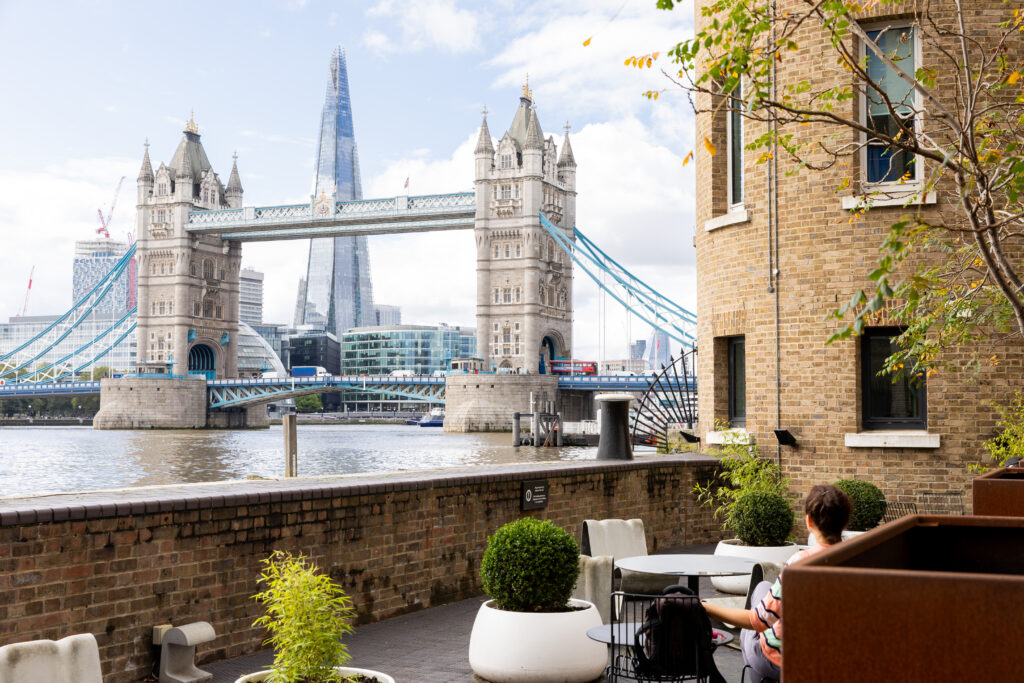 A person sits on a patio near the Thames River with Tower Bridge and the Shard visible in the background. The area is accented with potted plants against a brick building. The sky is partly cloudy.