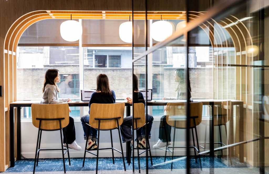 Three people sit at a high table with laptops in a modern, glass-walled office meeting room. The space features wooden accents, overhead round lights, and a view of buildings outside.
