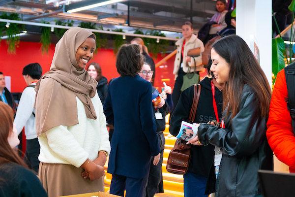 People engage in conversation at a lively event. A woman in a hijab smiles warmly at another woman showing her a phone. The background is a modern space with a red wall and plants, filled with other attendees.