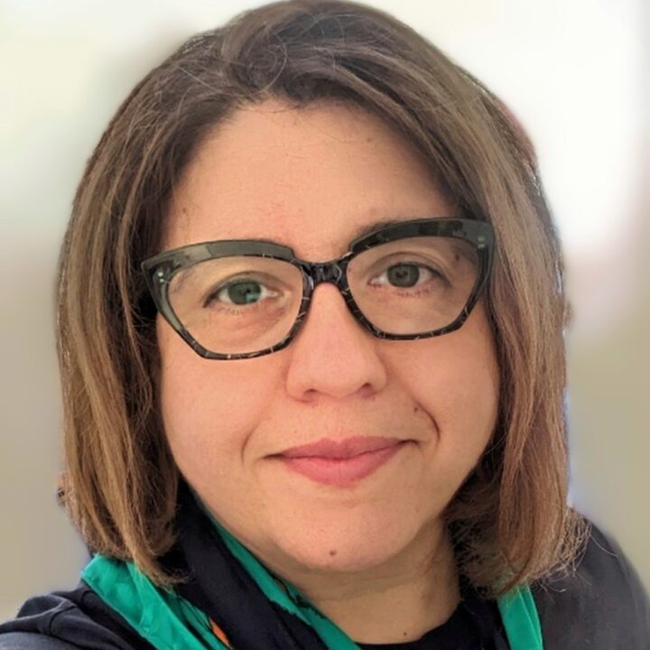 A person with shoulder-length brown hair, wearing black-rimmed glasses and a patterned scarf. The background is blurred. They are looking at the camera with a neutral expression.