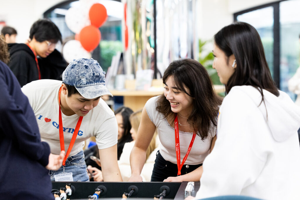Three people are playing foosball indoors. One person in a denim cap is bending over, focusing on the game. Two others, wearing red lanyards, are smiling and engaged. There are blurred figures and balloons in the background.