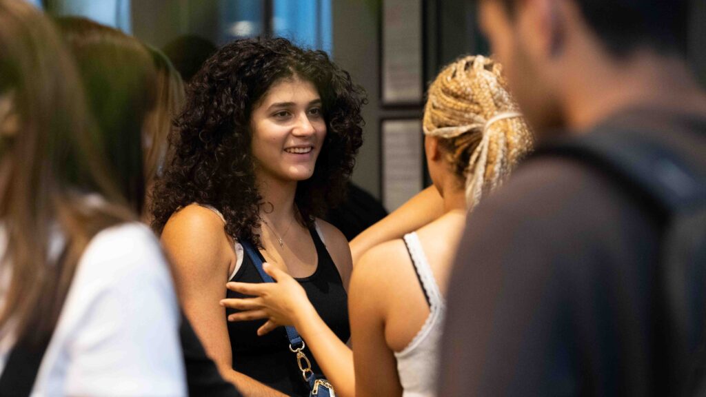 A young woman with curly hair and a black tank top smiles while talking to others in a casual indoor setting. People around her are engaged in conversation.