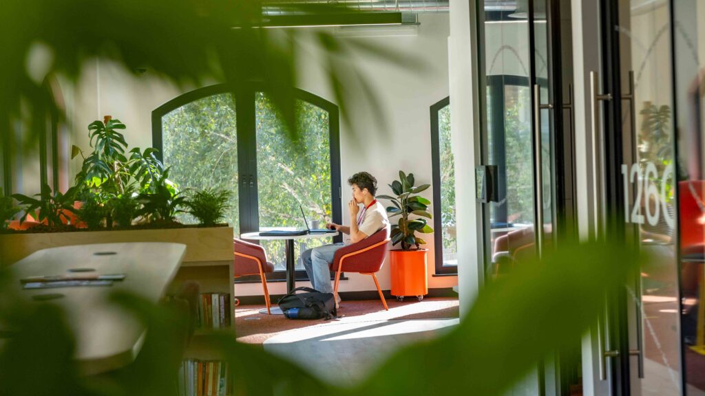 A person sits alone at a round table in a bright room with large windows, surrounded by green plants. They appear to be studying or working, with a backpack and a laptop on the table.